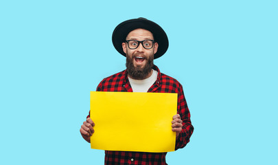 Handsome hipster man holding a blank yellow billboard or poster with blank space for your text isolated on blue background. Discount, sale, season sales.