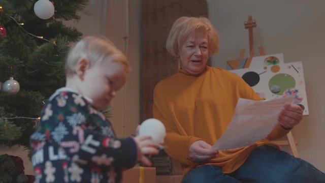 Adorable small baby child in sweater playing with toys at Christmas tree. Happy grandma unwrapping and admiring amazing painting paper. Family bonding.