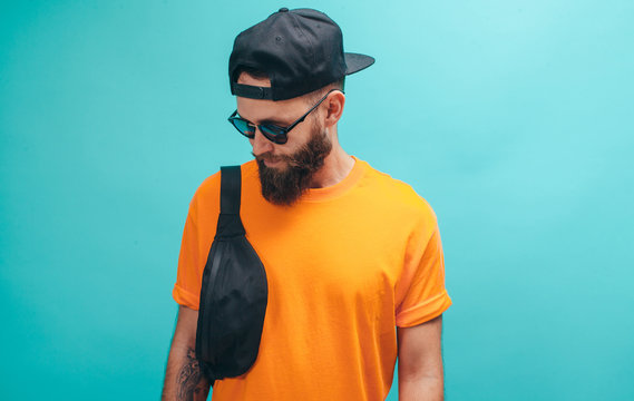 Handsome hipster guy with beard wearing orange tshirt, black waist bag and a cap on blue background. Mockup for print