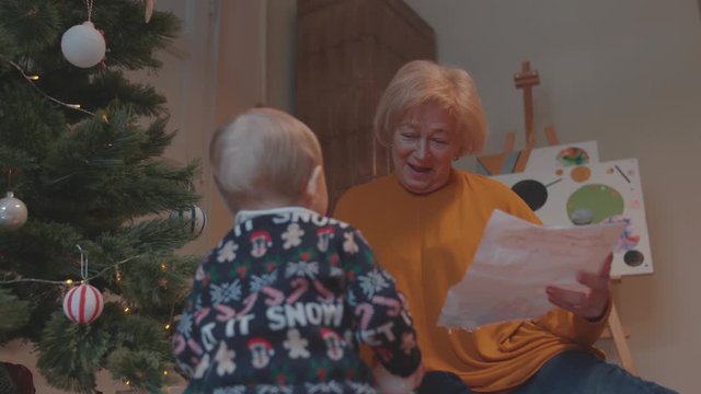 Joyful grandmother upbringing her cute grandchild baby playing and drawing together near Christmas tree at home. Winter family holidays.