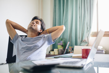 Young Asian office worker relaxing with hands behind head at office desk. Daydreaming concept