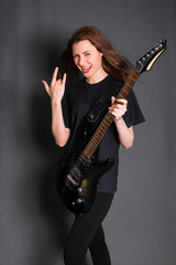 Naklejka premium A beautiful, young rock girl in black clothes with an electric guitar in her hands shows a goat gesture. Studio photo on a gray background. Model with clean skin.