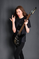 Fototapeta na wymiar A beautiful, young rock girl in black clothes with an electric guitar in her hands shows a goat gesture. Studio photo on a gray background. Model with clean skin.