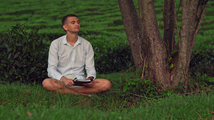 Naklejka na ściany i meble A young man with closed eyes sits on the grass under a tree with a notebook and enjoys nature.