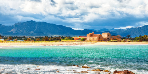 The Nora bay and beach, the medieval Sant'Efisio church near the shore and mountains in the background - obrazy, fototapety, plakaty