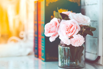 Beautiful artificial pink roses in glass vase on a white table with blured image of three books in...