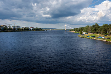 Obraz premium Republic of Finland, Rovaniemi: Kemijoki river next to the city center of the Finnish town with riverbank and modern white Jatkankynttila bridge (Lumberjack Candle Bridge) in background.