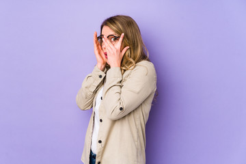 Young caucasian woman isolated on purple background blink through fingers frightened and nervous.