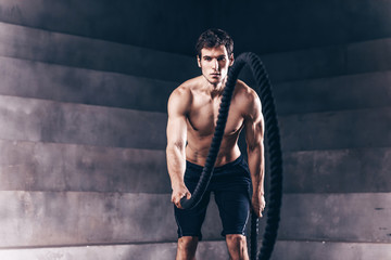 Handsome muscular man is doing battle rope exercise while working out in gym.