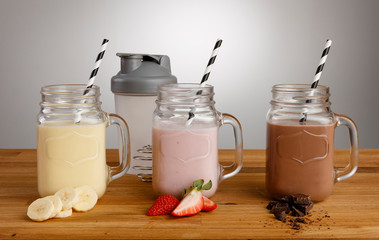 Banana, Strawberry and Chocolate milkshakes and ingredients in Mason Jar glasses, with paper straws and shaker on a wooden table top