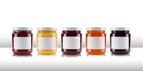 A group jars containing jams and preserves, including rasperry, strawberry, marmalade, chutney and relish, with blank white labels on a white table like surface