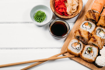 Japanese sushi rolls set with salmon acne and cream cheese on bamboo board with chopsticks and fresh food ingredients with soy sauce and ginger on white wood copyspace background.