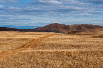 A road in the steppe going to the hills. Yellow grass. The weather is cloudy. Mountains in the distance.