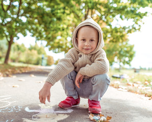 little boy, summer city, draws with crayons, casual warm clothes, beige sweater with hood. Creative creativity and art concept. Background autumn leaves asphalt. Emotions of happiness smile positive.