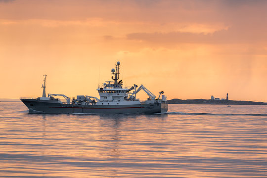 ULSTEINVIK, NORWAY - 2018 MAY 31. Fishing Vessel Passing Grasoyane Lighthouse In The Orange Sunset.