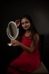 Fashion portrait of an young and attractive Indian Bengali brunette girl in red western dress holding a mirror in her hand in front of a black studio background. Indian lifestyle