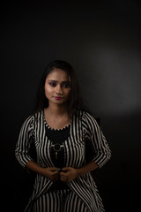 Fashion portrait of an young and attractive Indian Bengali brunette girl with striped western semi formal/casual dress in front of a black studio background. Indian fashion portrait and lifestyle.