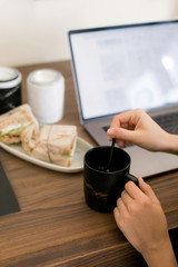 Fototapeta na wymiar The girl sitting at a working wooden table with a laptop decided to have a snack and eat a hot tea sandwich in a black mug. Lunch break. The gadgets and technology that surround us.