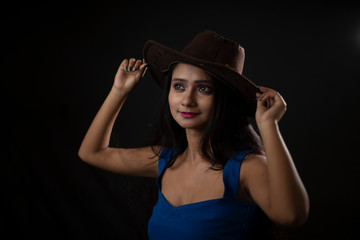 Fashion portrait of young and attractive Indian Bengali brunette girl with blue western dress with cowboy hat under spotlight in front of black studio background.Indian fashion portrait and lifestyle.