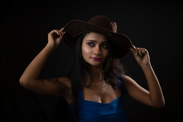 Fashion portrait of young and attractive Indian Bengali brunette girl with blue western dress with cowboy hat under spotlight in front of black studio background.Indian fashion portrait and lifestyle.