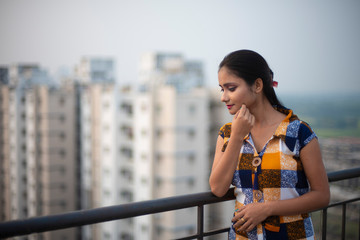 Fototapeta premium Fashion portrait of an young and attractive Indian Bengali brunette girl in western dress standing in a balcony/rooftop in urban background. Indian fashion portrait and lifestyle.