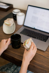 The girl sitting at a working wooden table with a laptop decided to have a snack and eat a hot tea sandwich in a black mug. Lunch break. The gadgets and technology that surround us.