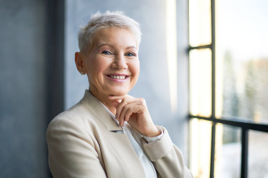 Happy middle aged blonde woman in stylish beige jacket standing by window in office, holding hand under her chin and smiling at camera. Success, business, people, lifestyle, age, work and employment