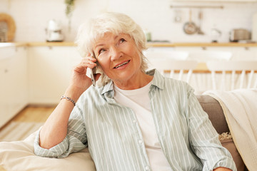 Indoor shot of charming friendly senior gray haired woman holding generic smart phone close to her ear, having hearing problem, talking to her friend, sitting comfortably on couch in living room