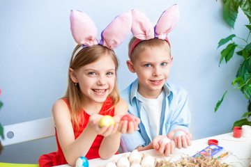 cute smiling children, fair-haired brother and sister of 7-9 years old, wear bunny ears on their heads and paint Easter eggs on a blue background