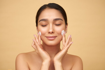 Young girl with closed eyes with clean skin puts cream on her face with hands on a yellow background