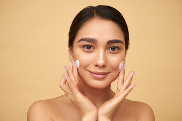 Young girl with clean skin puts cream on her face with hands on a yellow background