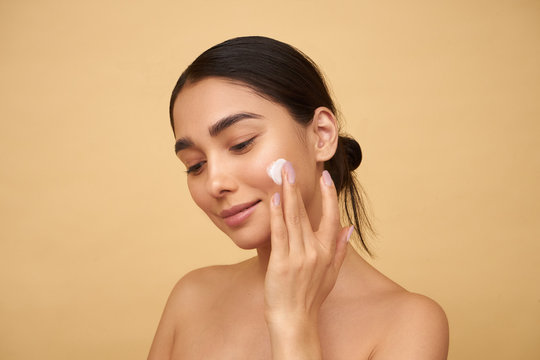 Young girl with clean skin applies cream on her face with one hand on a yellow background