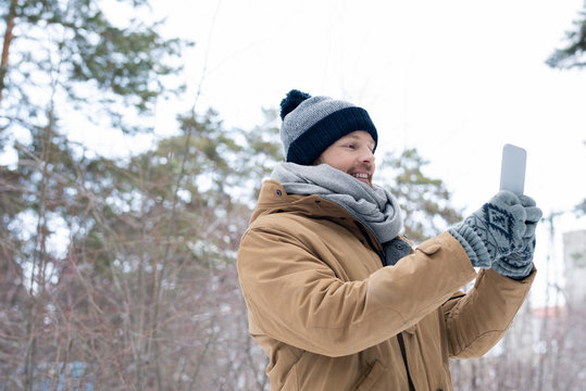 Horizontal Low Angle Portrait Of Young Man Wearing Casual Warm Outfit Taking Selfie Shot On Smartphone Camera On Winter Day