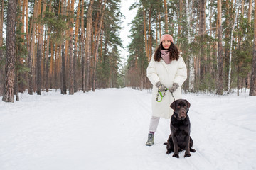 Fototapeta na wymiar Horizontal shot of stylish young woman in warm winter outfit standing on urban forest road with chocolate labrador retriever