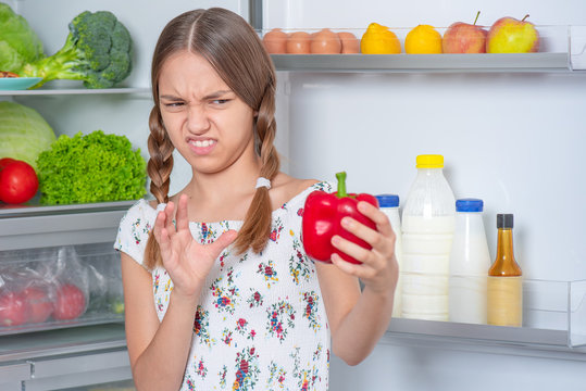 Teen girl holding fresh red pepper while standing near open fridge in kitchen at home. Child do not want to eat vegetables and dislike taste of pepper. Organic natural healthy food produce.
