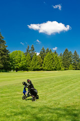 A golf bag on three wheel golf cart over a beautiful green with blue sky and white clouds.