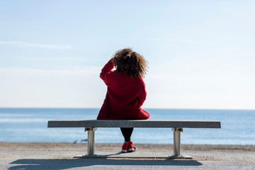 Rear view of a young curly woman wearing red denim jacket sitting on a bench while looking away to...