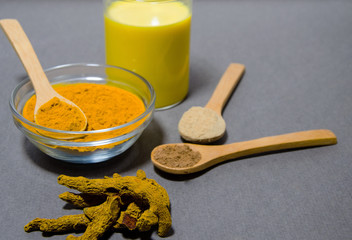 Turmeric root lies on a gray background. Nearby is a glass bowl with ground turmeric and a wooden spoon, ground ginger and cinnamon in wooden spoons lie nearby. In the background is a glass of turmeri