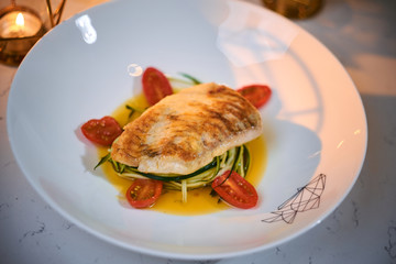 Delicious fillet of grilled and oven baked codfish or coalfish served with thin sliced vegetables and oil sauce, on a white porcelain dish next to golden candles in a warm light of a restaurant, close