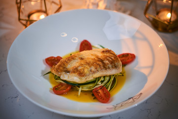 Delicious fillet of grilled and oven baked codfish or coalfish served with thin sliced vegetables and oil sauce, on a white porcelain dish next to golden candles in a warm light of a restaurant, close