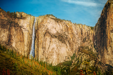 Waterfall in Yosemite national park, California, USA. Scenic landscape with waterfall, stone rocks, sequoia trees in sunny day with blue sky above. Travel tourist destination in CA