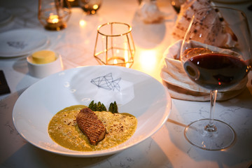 Haute cuisine fillet of grilled and oven baked gourmet fish served with garnish including herbs and oil sauce, on a white porcelain dish next to golden candle, a glass of red wine and a full wicker