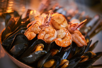 A full dinner rich ceramic bowl of prepared grilled shrimps and mussels including tiny octopus served on a white stone table next to golden candle in a soft light of a restaurant, close up