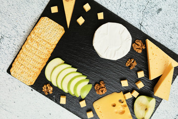 Pieces of cheese on a dark background. Cheeseboard. Sliced ​​apple and nuts on the board. Hard cheese, camembert, roquefort, emmental.