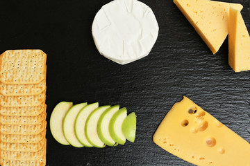 Pieces of cheese on a dark background. Cheeseboard. Sliced ​​apple and nuts on the board. Hard cheese, camembert, roquefort, emmental.