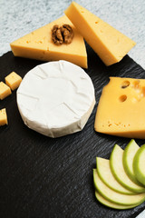 Pieces of cheese on a dark background. Cheeseboard. Sliced ​​apple and nuts on the board. Hard cheese, camembert, roquefort, emmental.
