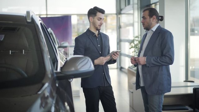 Side view of confident adult Caucasian businessman talking with young car dealer in showroom. Two men standing in dealership and discussing purchase of automobile. Business, industry, lifestyle.