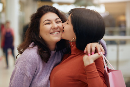 Сlose up portrait of two beautiful young women meeting in shopping center, brunette girl kissing her friend on cheek, lady dresses purple sweater holding paper bags with purchases, express happyness.