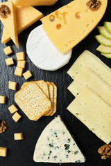 Pieces of cheese on a dark background. Cheeseboard. Sliced ​​apple and nuts on the board. Hard cheese, camembert, roquefort, emmental.
