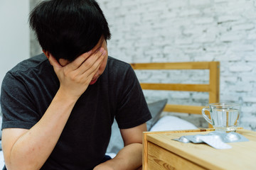 Obraz premium Young Asian man feeling sick and taking pills while sitting on the bed at home with medicines tablets and drinking water on table. Healthcare and Medicine concept